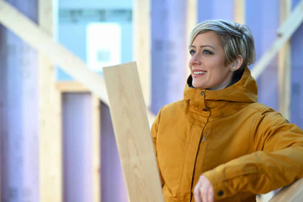 Erin Helland founder of HardHat Diplomat stands in wooden framework of a new addition being built. She is wearing a yellow midweight jacket