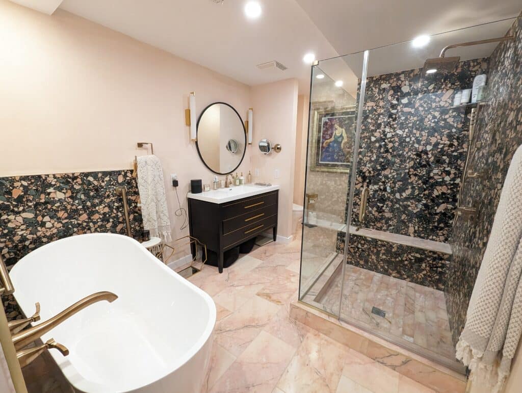 A newly designed bathroom in Washington, D.C., featuring a standalone white bathtub with brass fixtures, next to a unique terrazzo-like black, pink, and green tiled wall. A glass-enclosed shower with the same tile design, a modern vanity with a black base and brass handles, a circular mirror above it, and marble pink flooring complete the luxurious look. The room is well-lit with soft overhead lights, creating a warm ambiance
