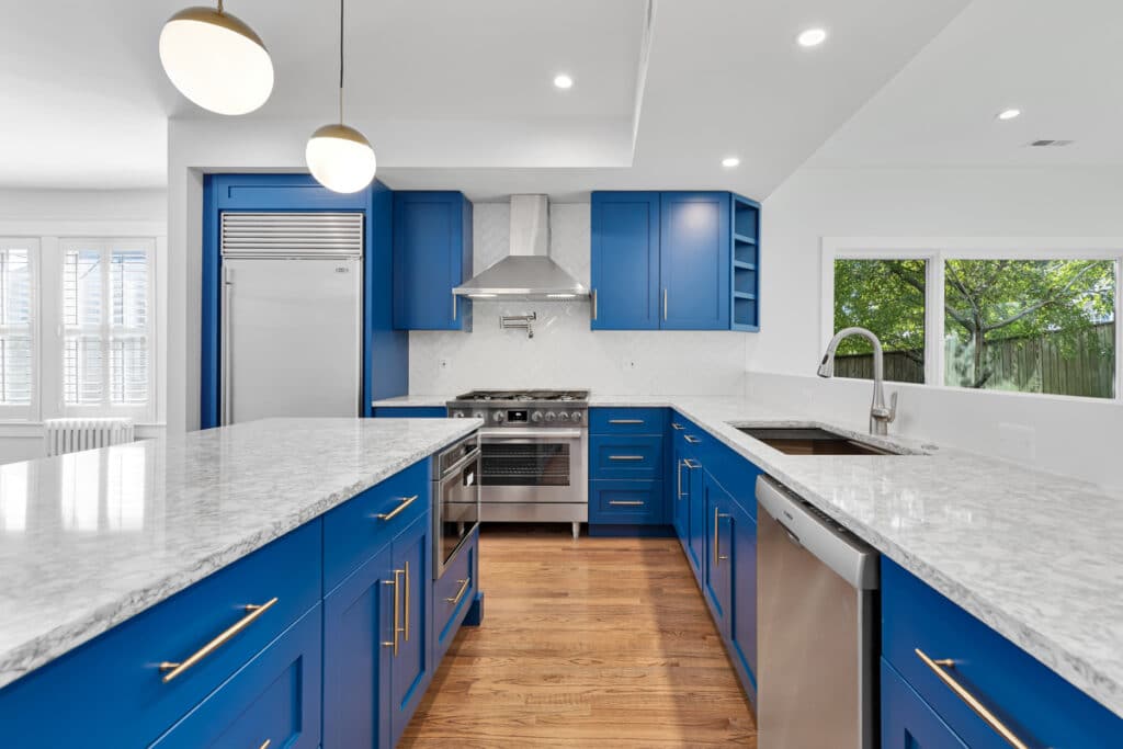 A modern kitchen in Washington, D.C., with vibrant blue cabinets, stainless steel appliances, and white countertops with a marble pattern. The kitchen features a gas range stove and a large refrigerator, with herringbone patterned white backsplash. Natural light streams in from windows with plantation shutters, highlighting the warm wooden floor.